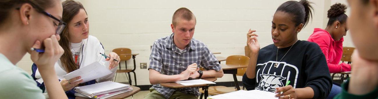 Five students sit with their desks pushed together discussing an assignment.