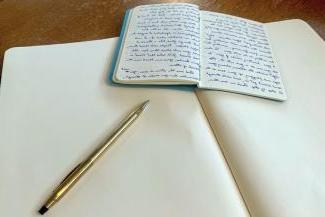 A notebook with writing lays on top of a blank notebook by a pen.